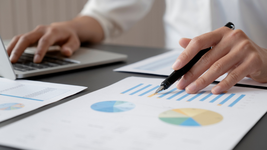 A close-up view of a person wearing a white shirt working at a dark gray desk. Their left hand is typing on a silver laptop, while their right hand holds a black fountain pen, pointing at a document filled with colorful bar charts and pie charts. Additional data reports are scattered on the desk.
