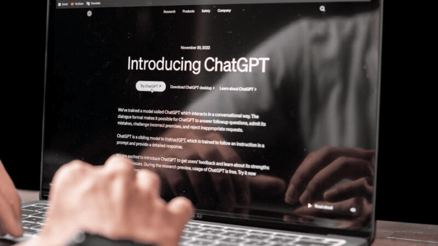 Gros plan sur un écran d'ordinateur portable affichant la page de présentation « Introducing ChatGPT » sur le site d'OpenAI, datée du 30 novembre 2022. Au premier plan, les mains d'une personne sont en train de taper sur le clavier, et le reflet de l'utilisateur est visible sur l'écran brillant de l'ordinateur.