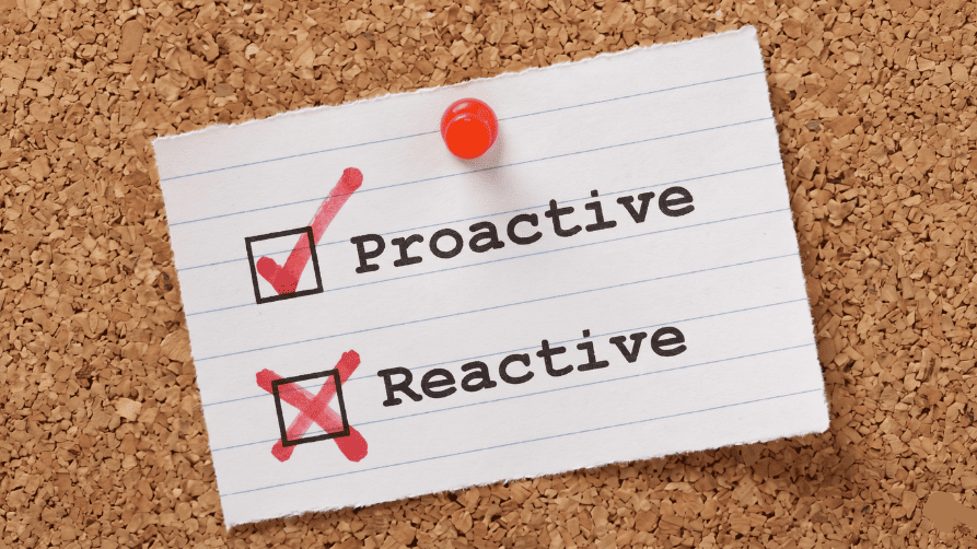 A close-up of a lined paper note pinned to a corkboard with a red pushpin. The note has two items listed with checkboxes. The first item, "Proactive," has a red checkmark in its box. The second item below it, "Reactive," has a red 'X' marked over its box.