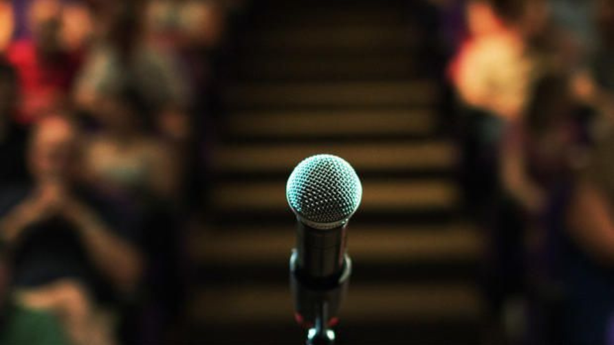 A sharp close-up shot of a metallic vocal microphone on a stand, positioned centrally on a stage. In the soft-focus background, rows of audience members sit on bleachers or auditorium stairs under ambient lighting.
