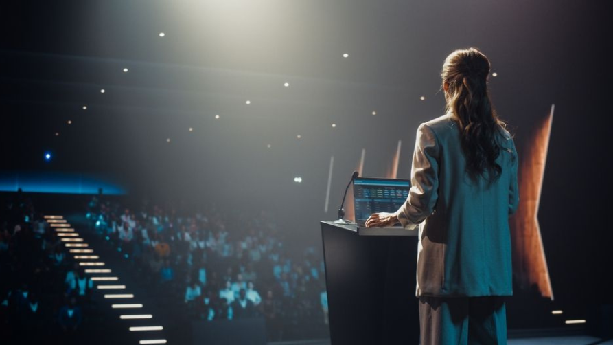 A woman with long wavy hair, wearing a professional suit, stands at a podium with a laptop and microphone under bright stage lights. She faces away from the camera toward a crowded, darkened auditorium.
